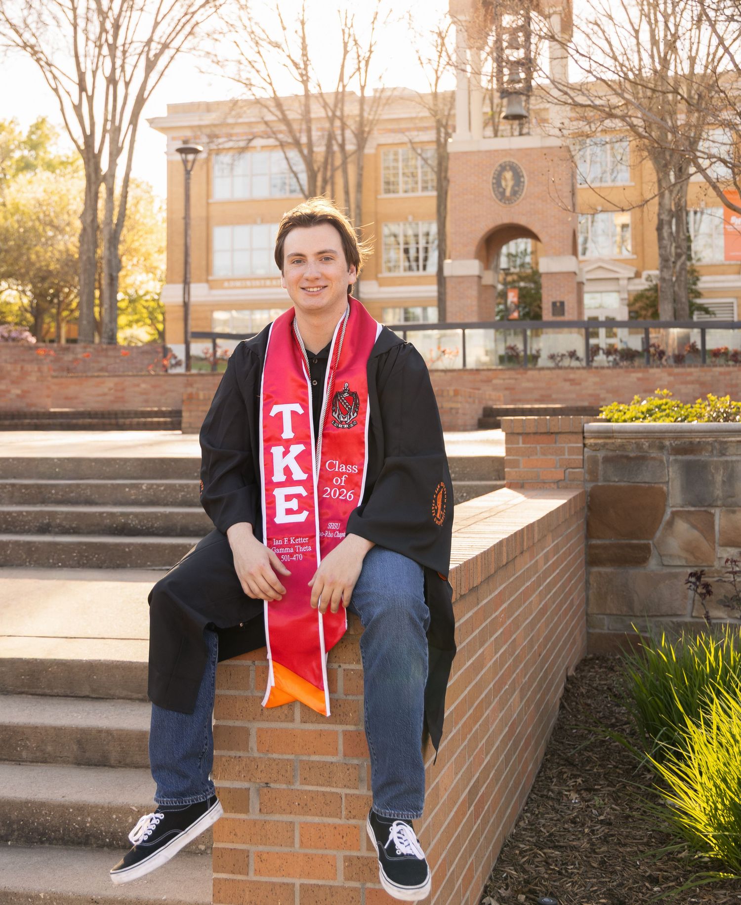 Male graduate in a black gown and red TKE stole sits on a brick ledge with a campus clock tower behind him in warm sunlight.