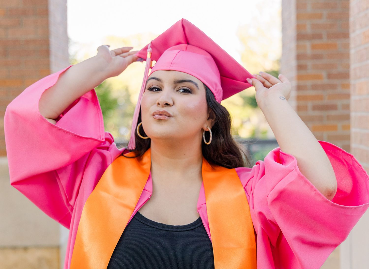 Graduate in a bright pink cap and gown with an orange stole poses playfully outdoors, adjusting her cap between brick columns.