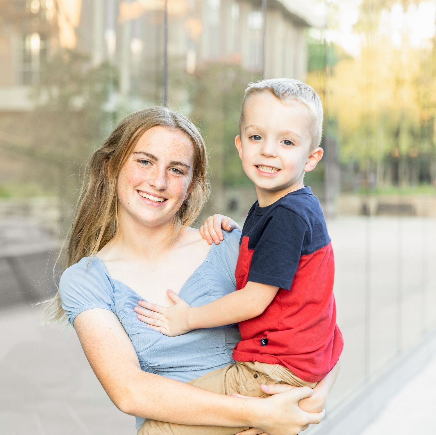 Smiling young woman in a blue top holding a happy little boy in a red and navy shirt outside near glass buildings.