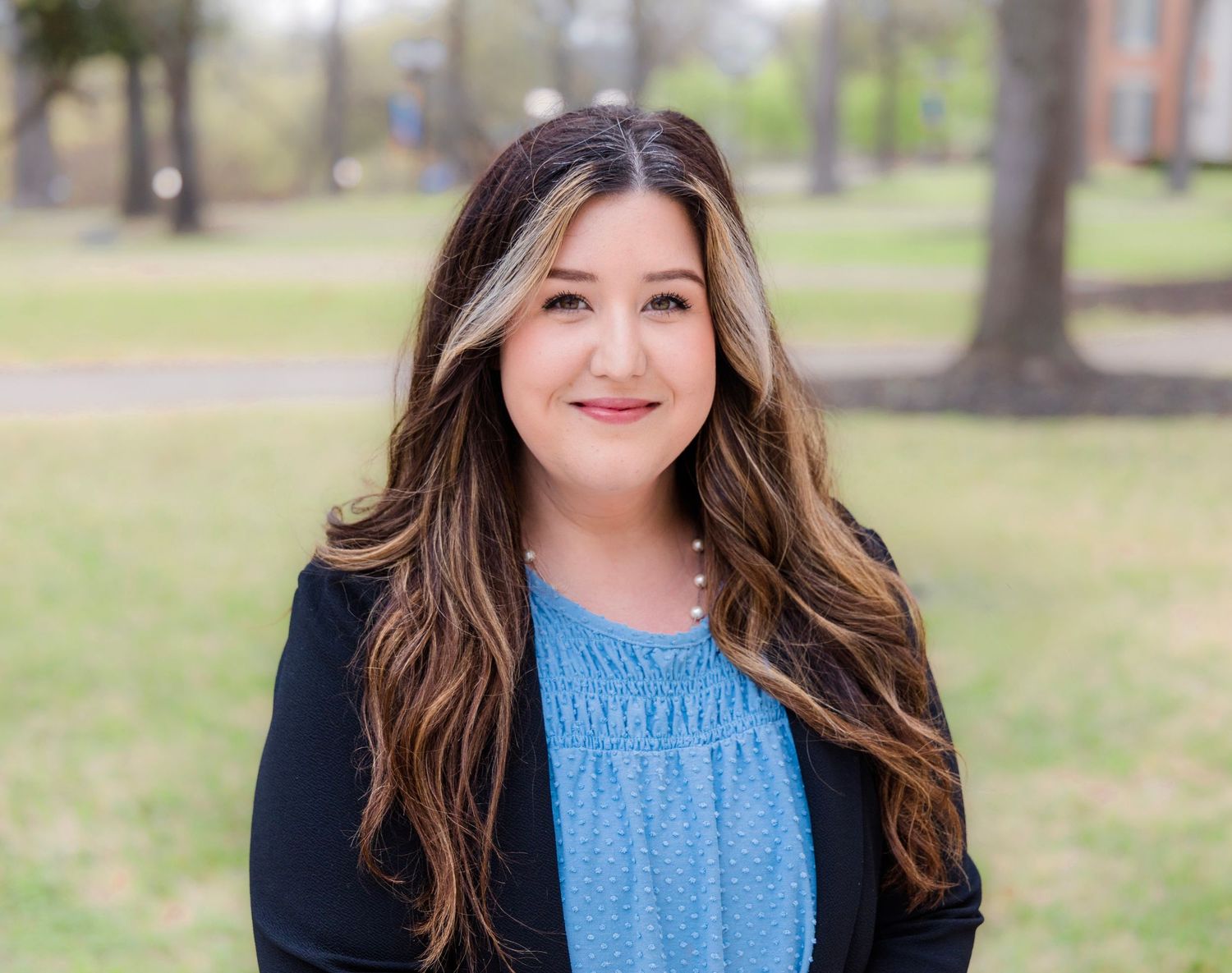 Smiling woman with long highlighted brown hair wearing a blue blouse and black blazer poses outdoors in a park with tall trees.