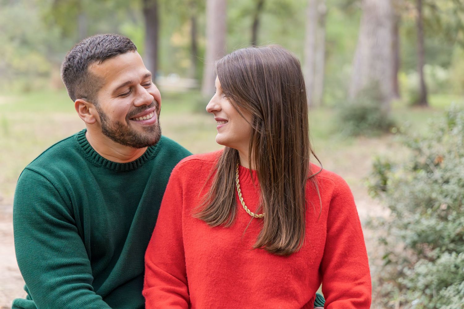 Smiling man in a green sweater and woman in a red sweater sitting close together in a sunny wooded park.
