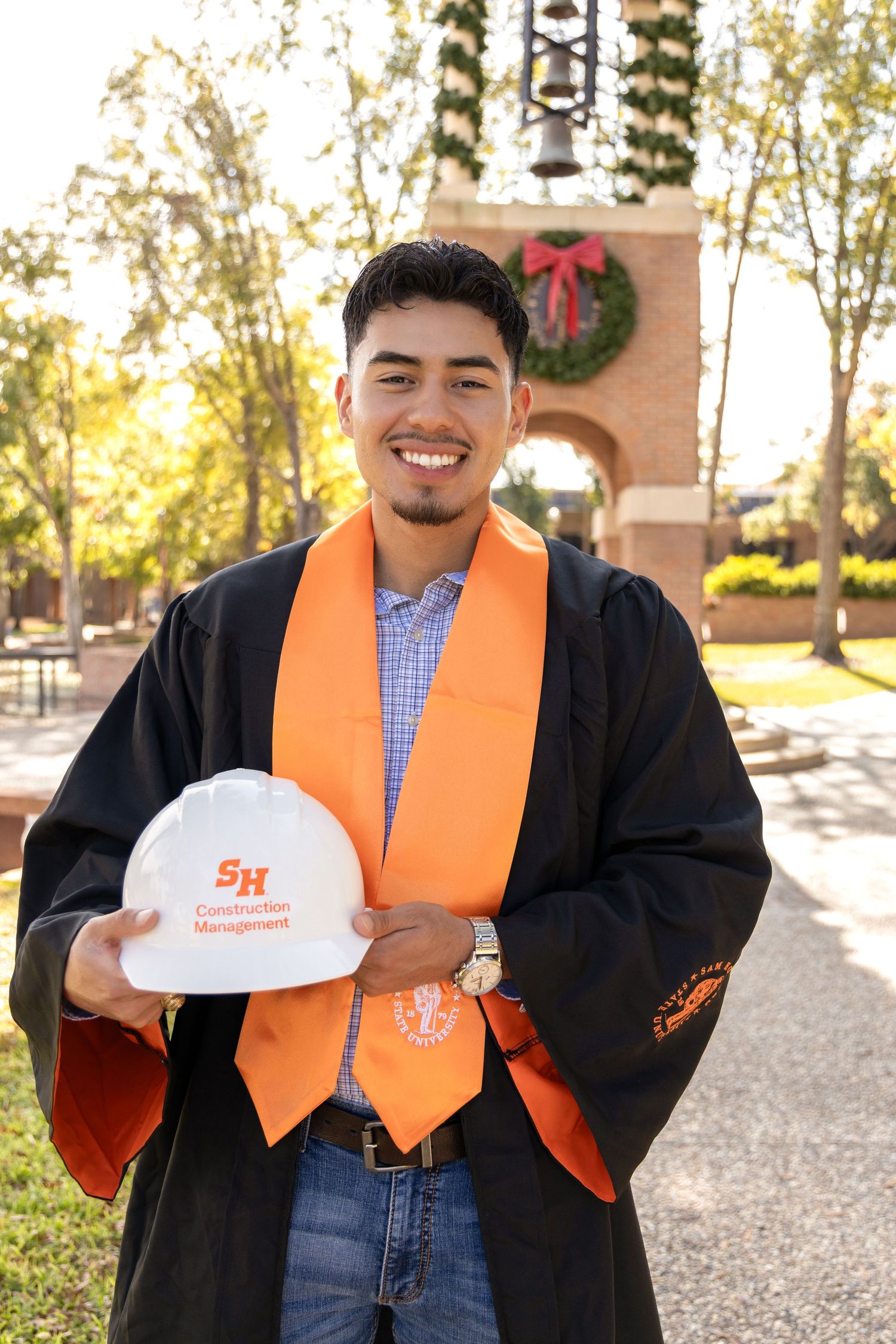 Smiling graduate in a black gown and orange stole holds a white Construction Management hard hat in front of a brick campus archway.