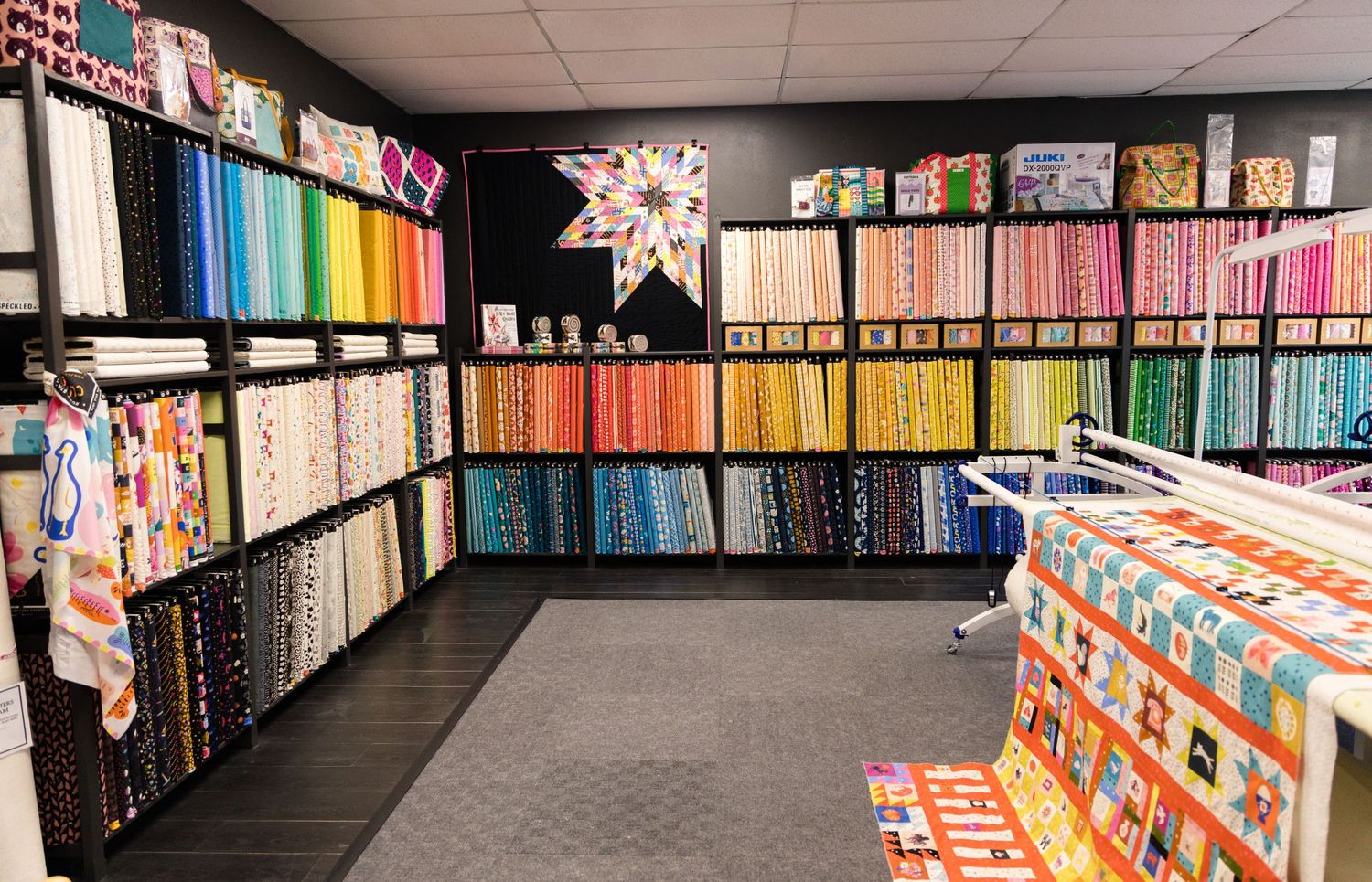 Interior of a fabric and quilt shop with shelves of colorful fabric bolts, a hanging star quilt and a quilt-covered table.