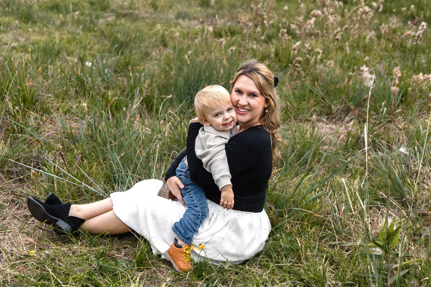 Smiling woman in a white skirt sits in a grassy field holding a toddler in a gray sweater and blue pants.