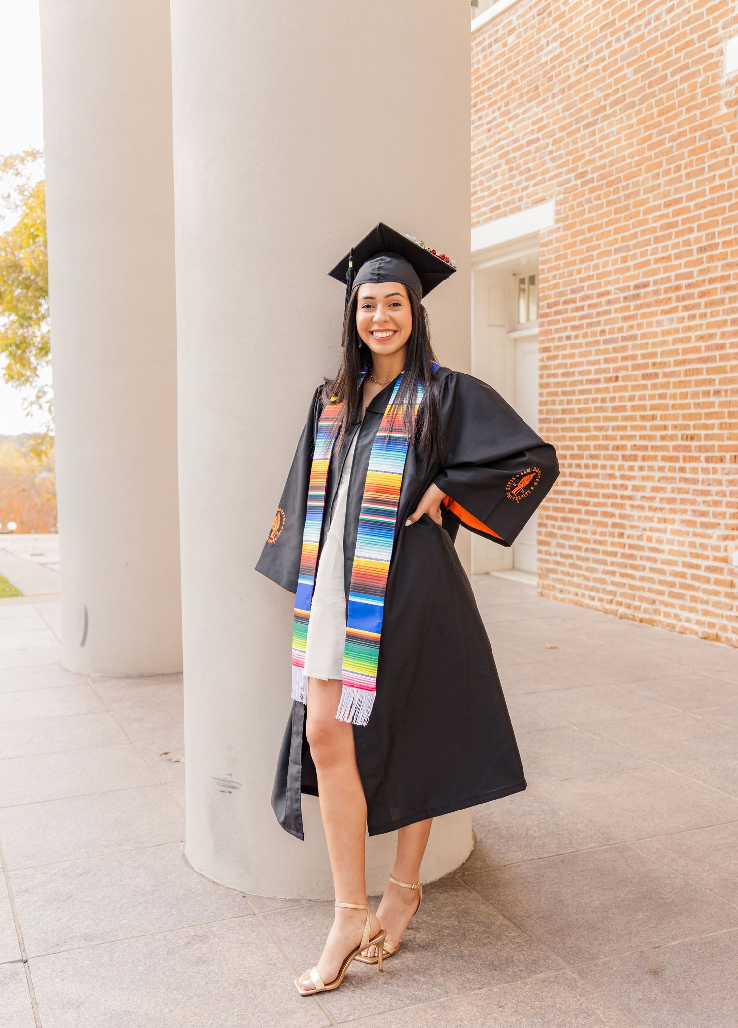 Smiling graduate in a black cap and gown wears a colorful striped serape stole while leaning against a white column by a brick building.