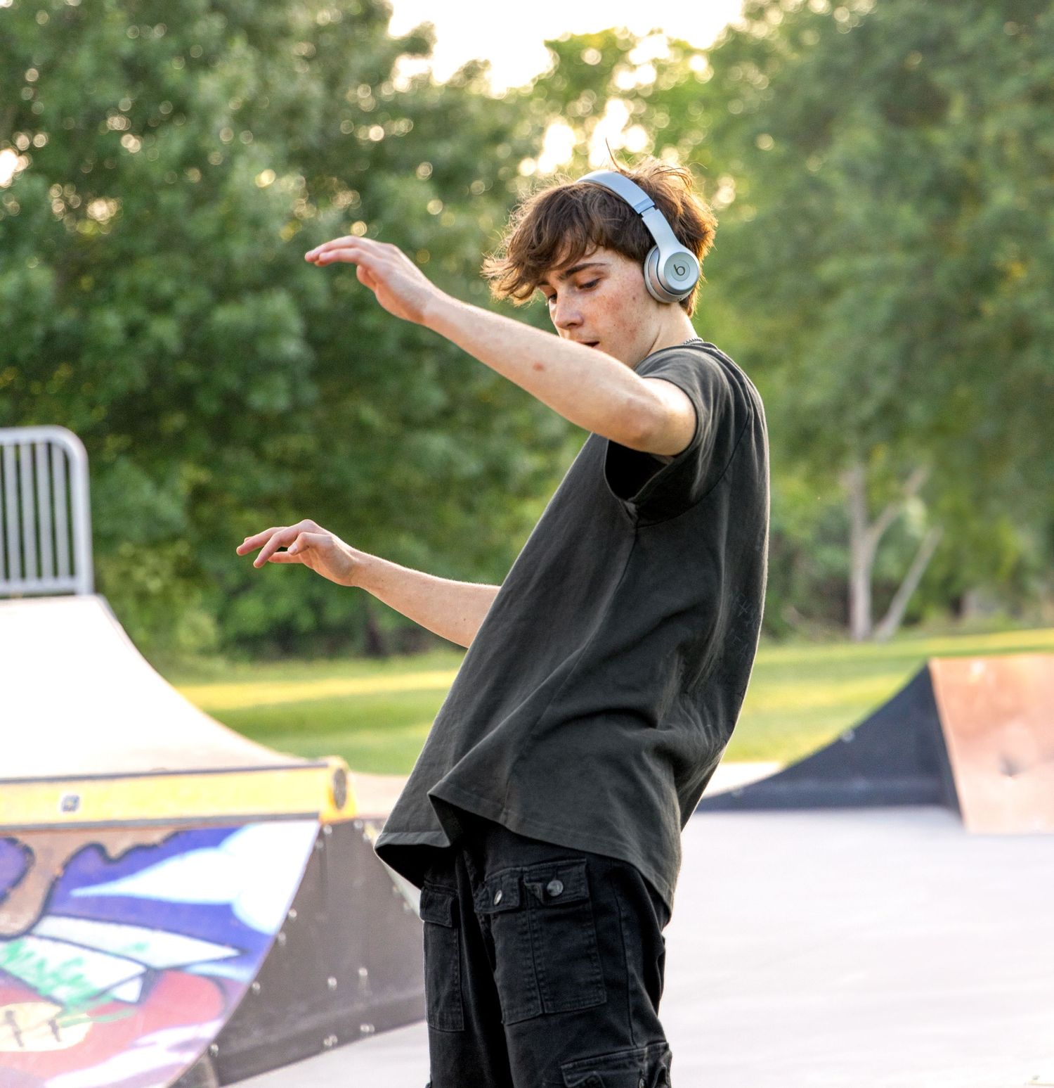 Teen wearing headphones and a black t-shirt dances with arms outstretched at a colorful skate park surrounded by green trees.