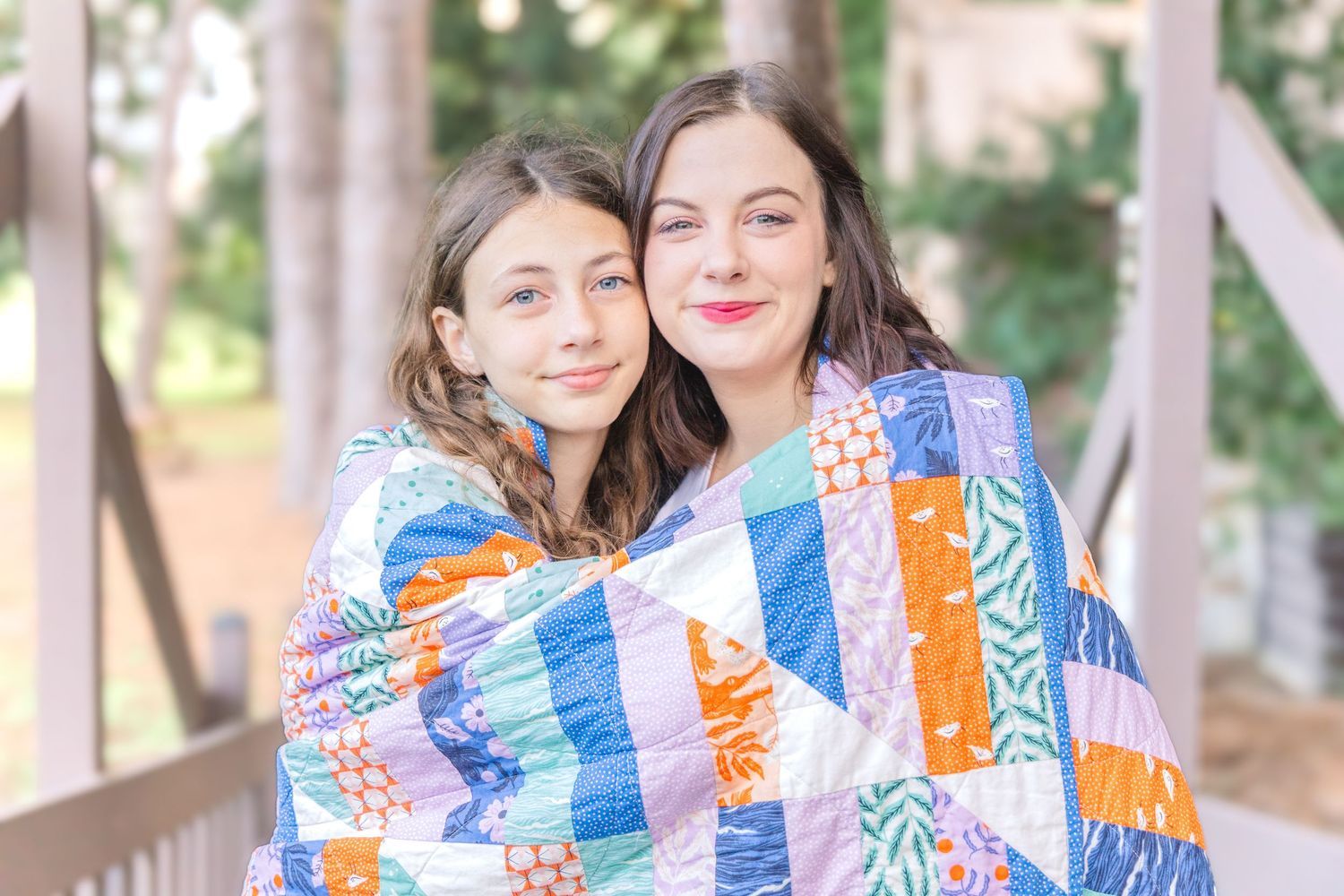 Two smiling young women snuggle together wrapped in a colorful patchwork quilt on a wooden porch with trees behind them.
