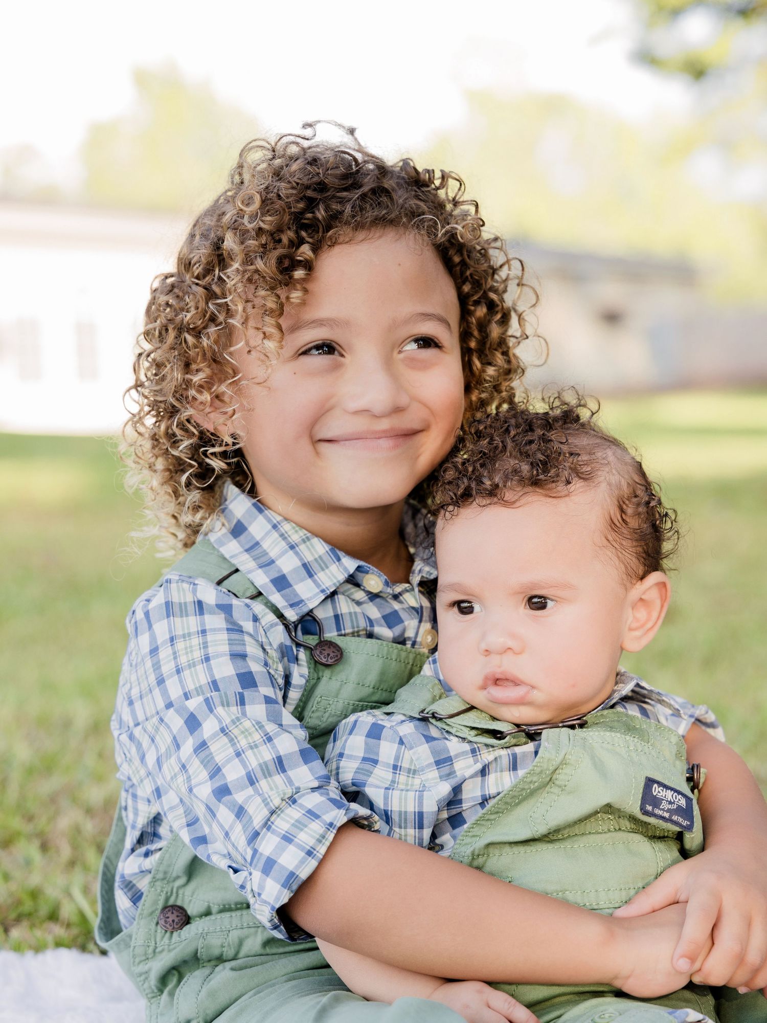 Young curly-haired child in a plaid shirt and green overalls hugging a baby in a matching outfit on a grassy lawn.