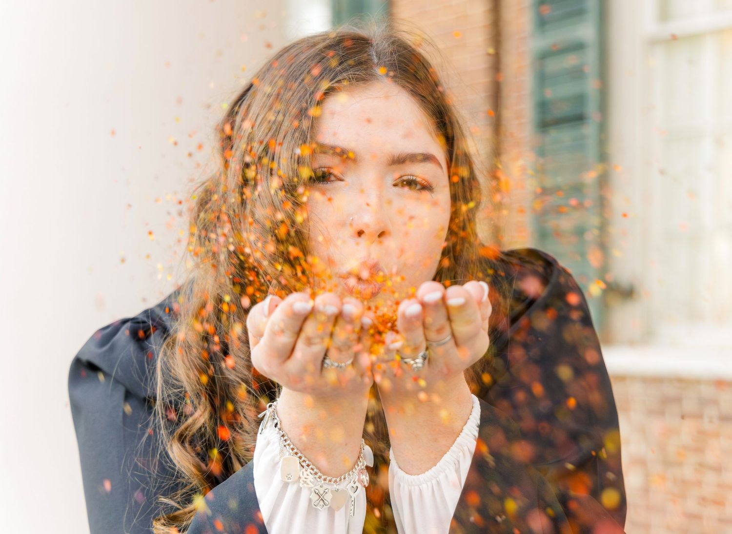 Young woman in a dark jacket blows orange glitter toward the camera, creating a festive sparkling effect in front of a brick building.