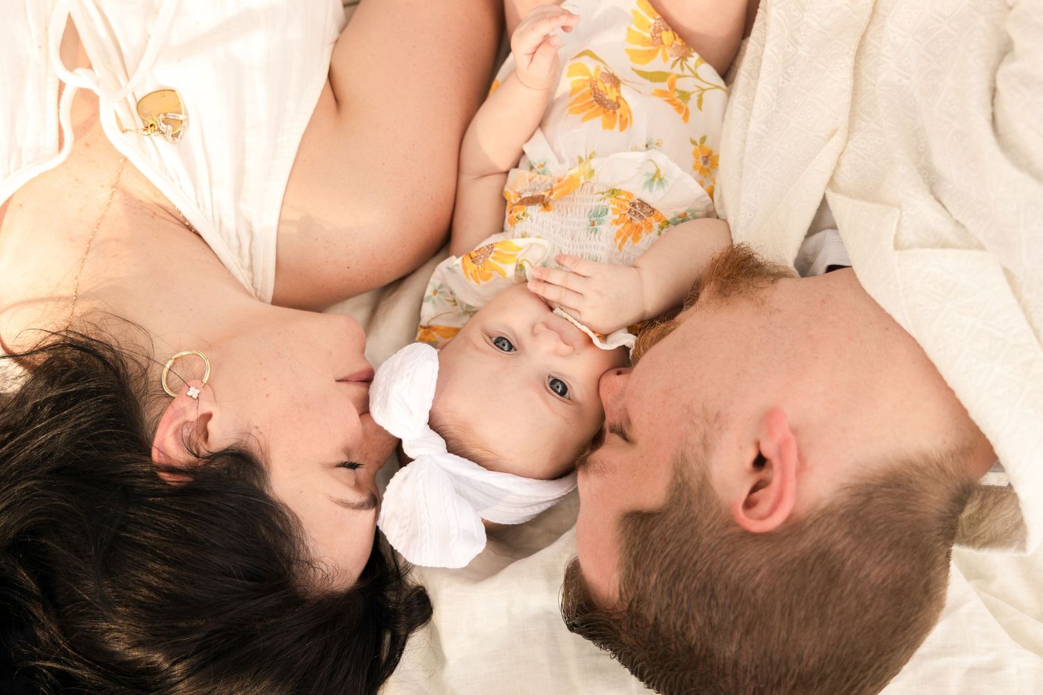 A mother and father lie side by side kissing their smiling baby girl in a floral romper and white bow on soft bedding.