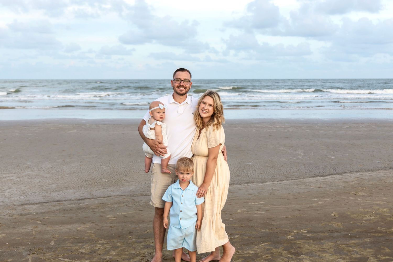 Smiling family of four with a man, woman, baby, and young boy pose together on a beach with ocean waves behind them.