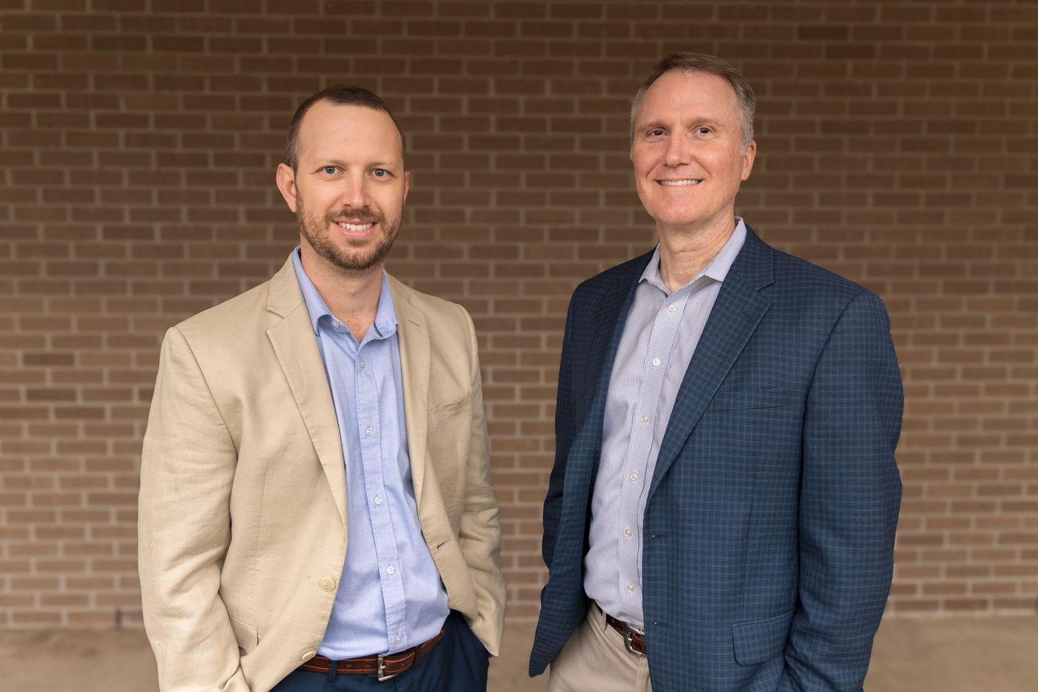 Two smiling men in blazers, one tan and one navy, stand side by side in front of a brick wall for a professional portrait.