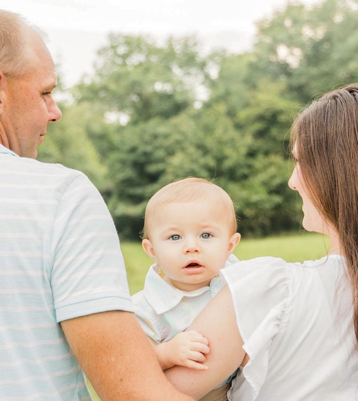 Smiling baby in a white collared outfit looks at the camera while held by two adults outdoors in front of green trees.