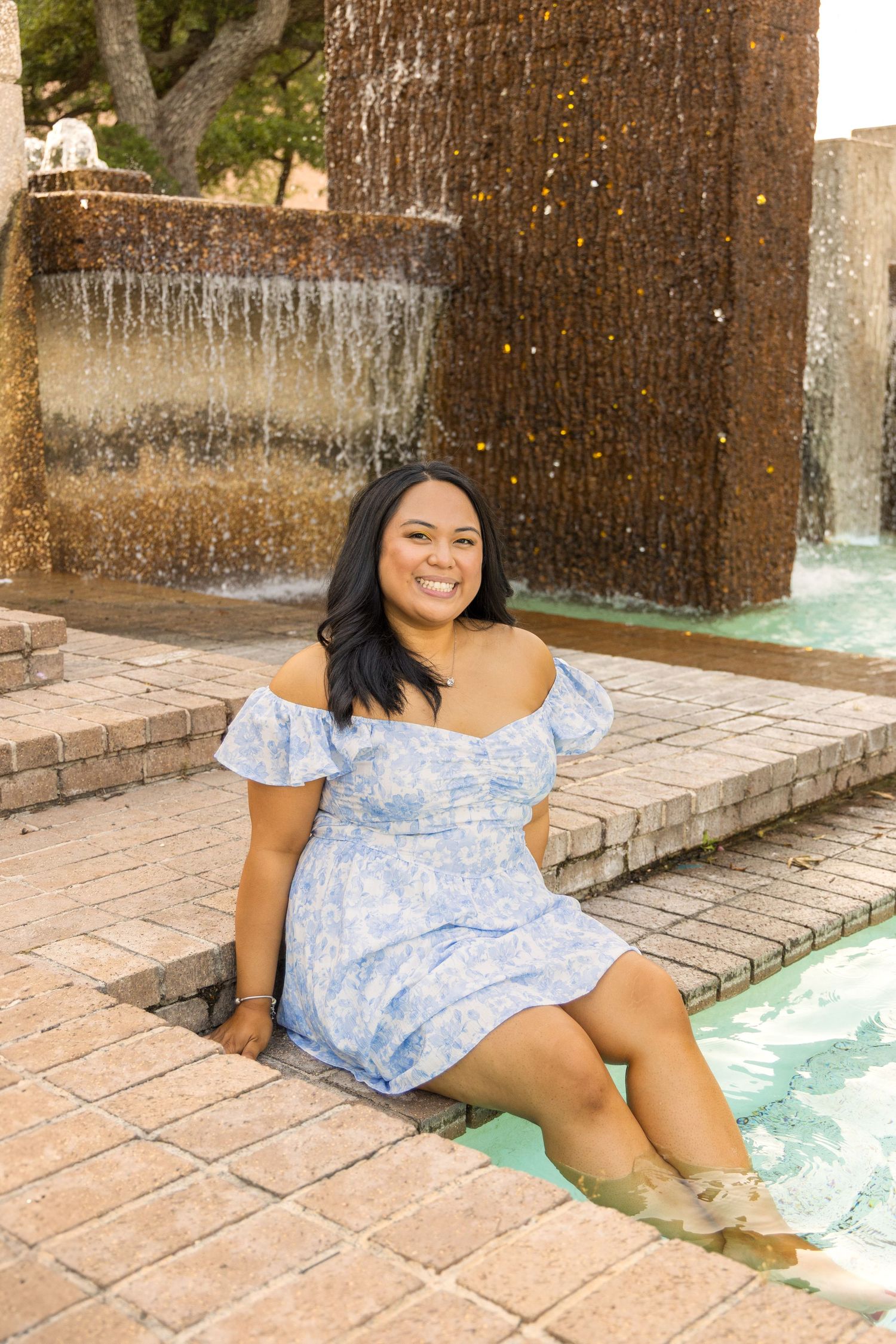 Smiling woman in a blue floral off-the-shoulder dress sits on brick steps with her feet in a campus fountain pool.