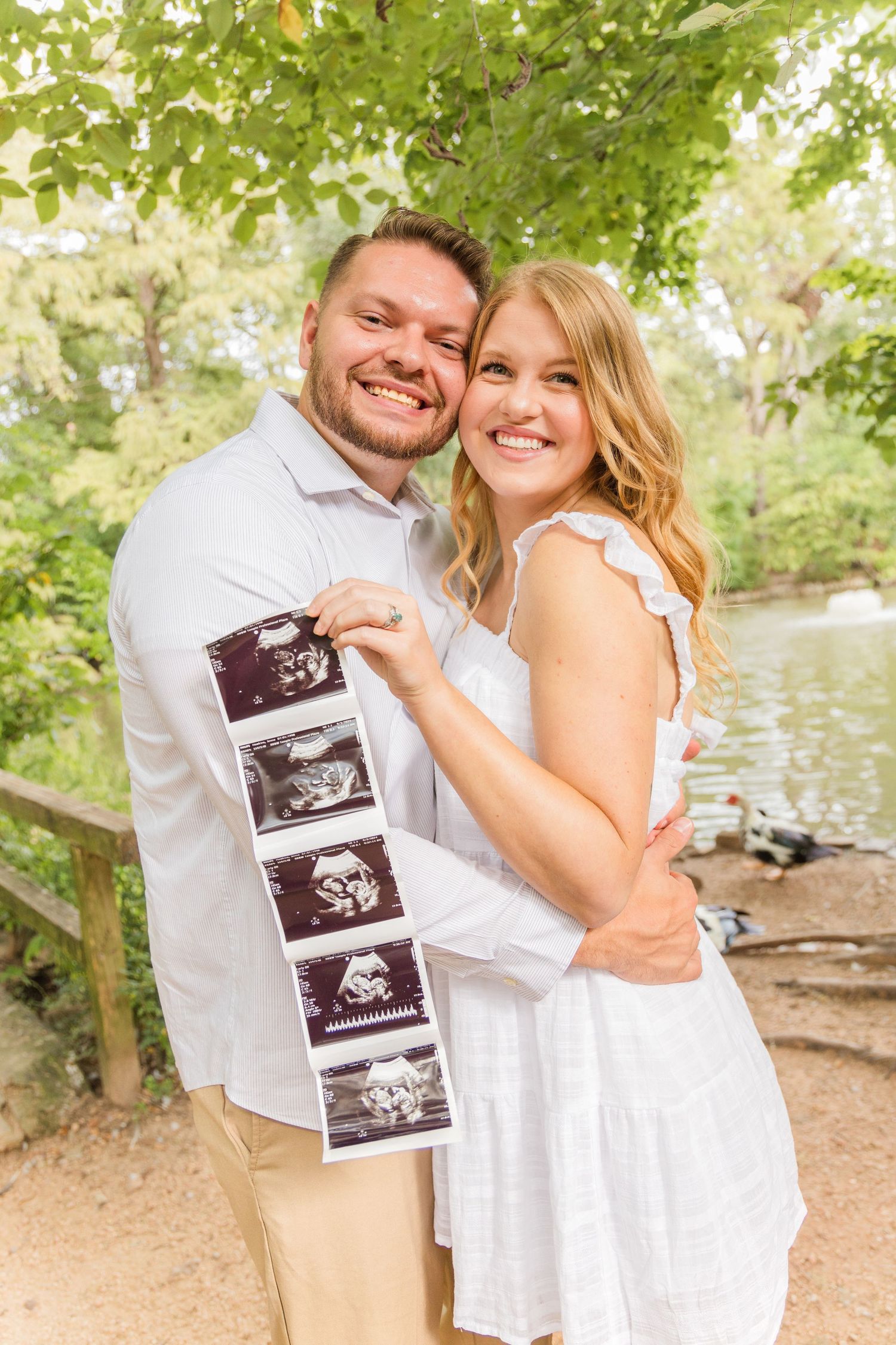 Smiling couple embracing outdoors by a pond while holding a strip of ultrasound photos to announce their pregnancy.