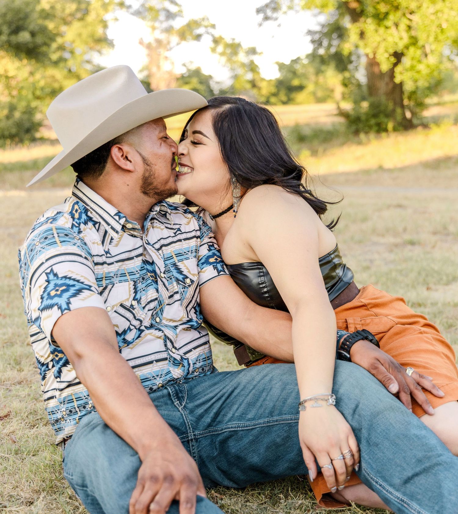 Couple in western attire shares a sweet kiss while sitting in a sunlit grassy field, the man wearing a cowboy hat and patterned shirt.