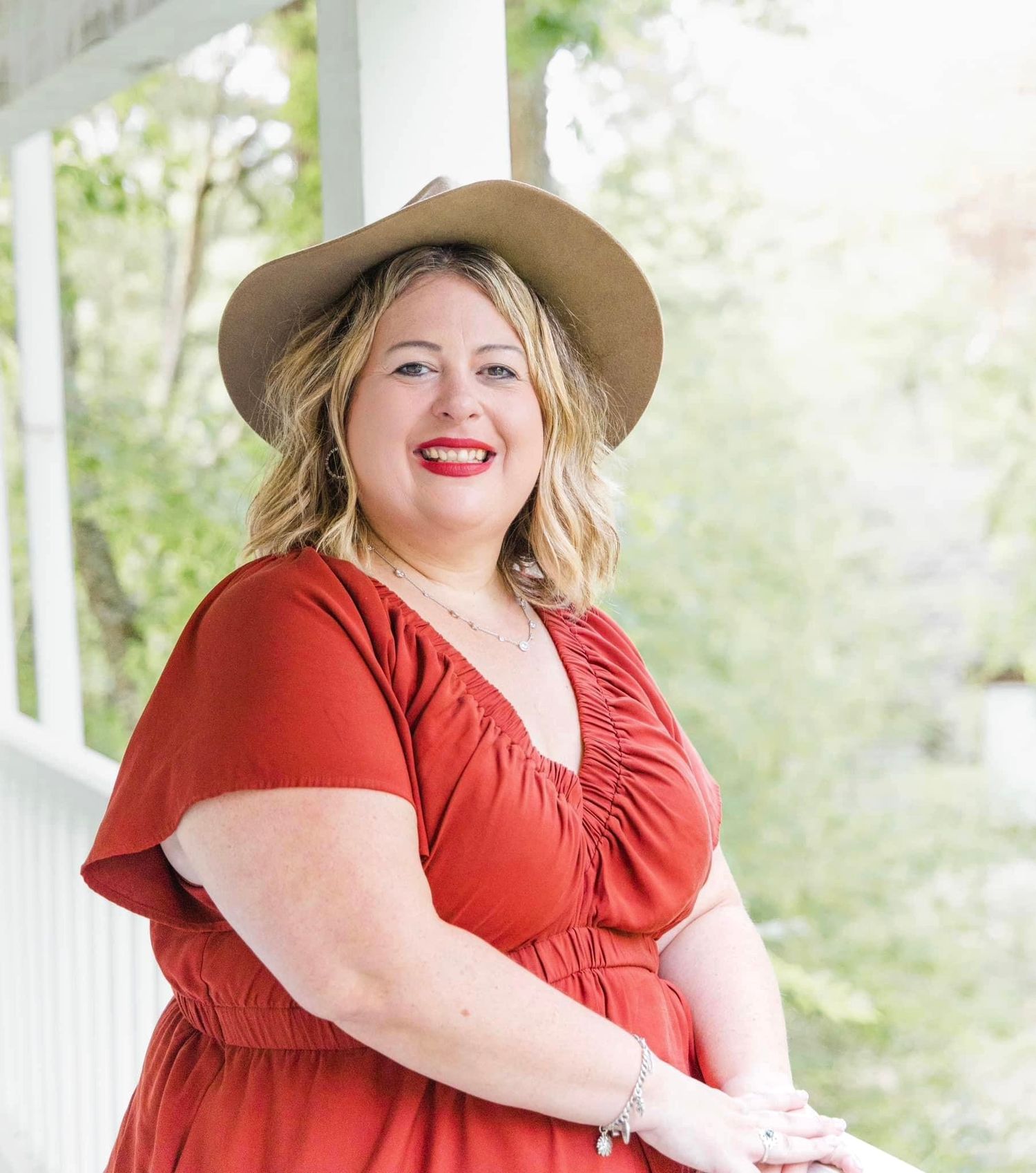 Smiling woman in a rust-colored dress and tan wide-brim hat with red lipstick poses on a white porch with green foliage behind.