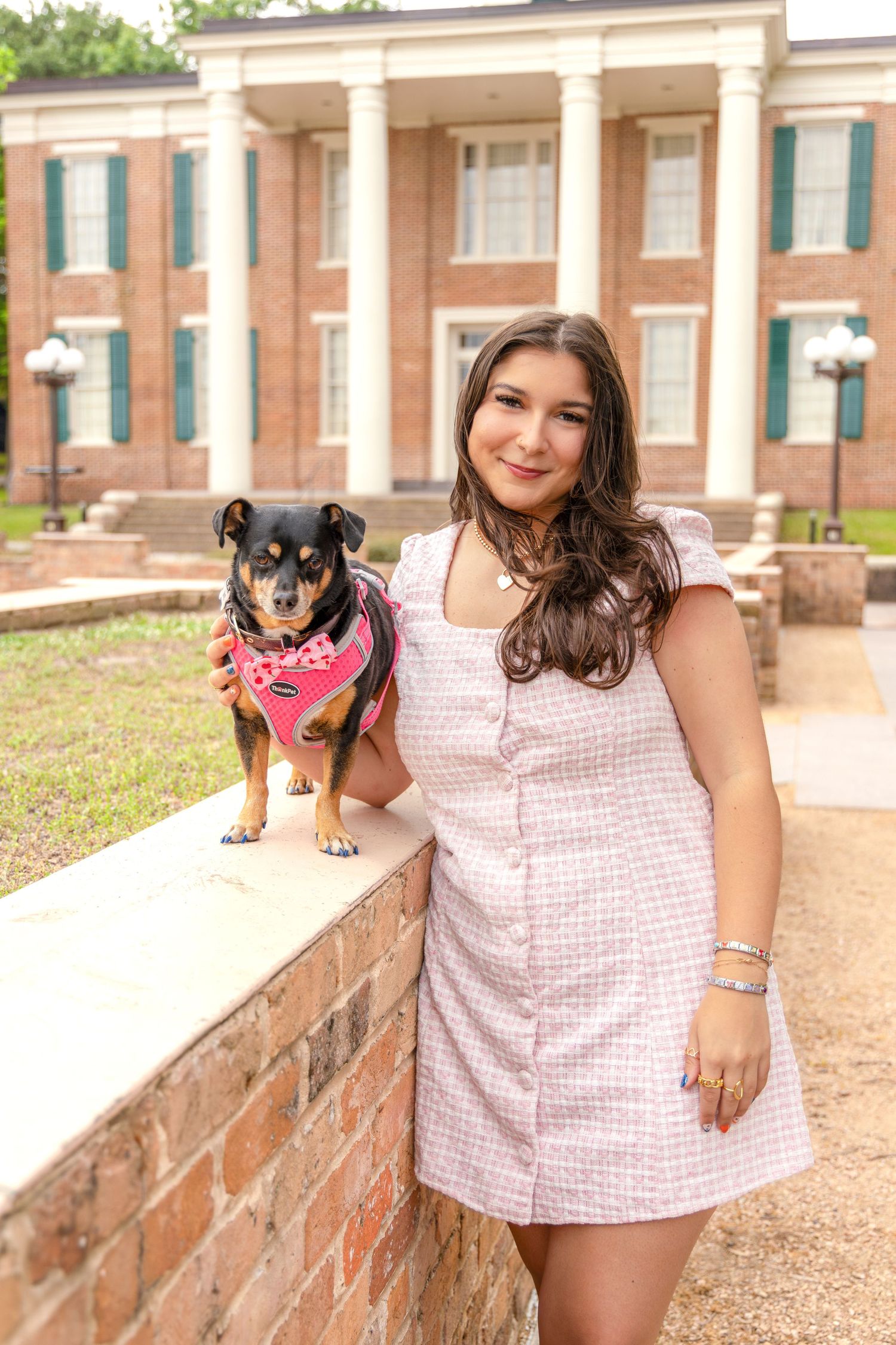 Woman in a pink tweed dress stands beside a small dog in a pink harness on a brick wall in front of a historic building.