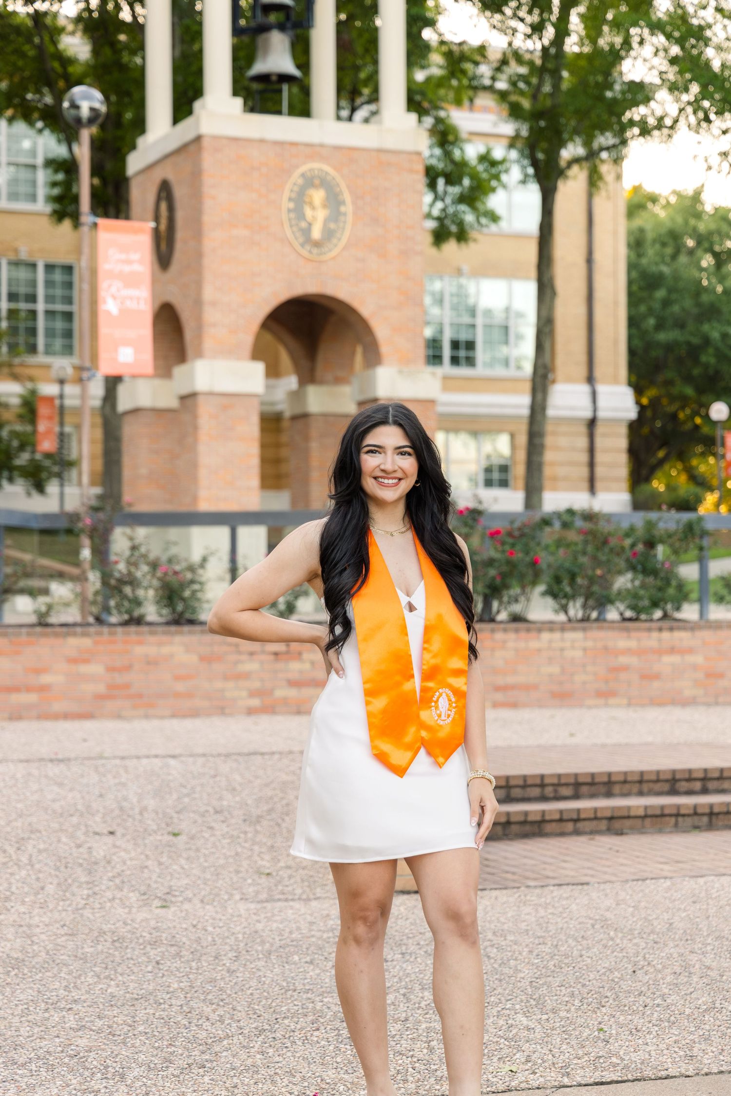 Smiling graduate in a white dress and orange stole poses on a campus walkway with a brick bell tower in the background.