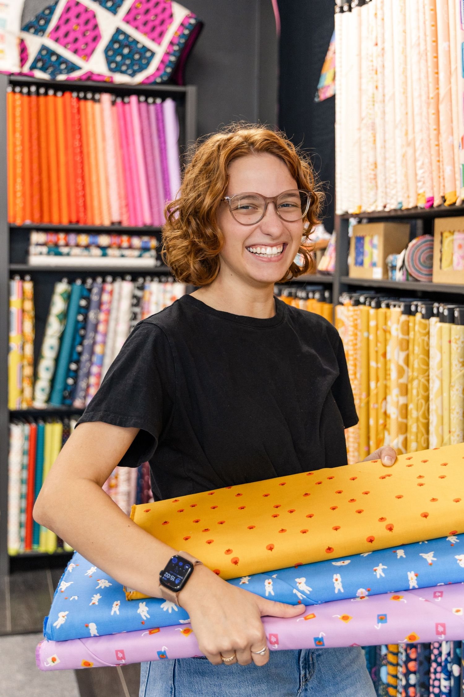 Smiling woman with glasses and curly red hair holds folded colorful fabric bolts inside a quilting shop with shelves of fabric.