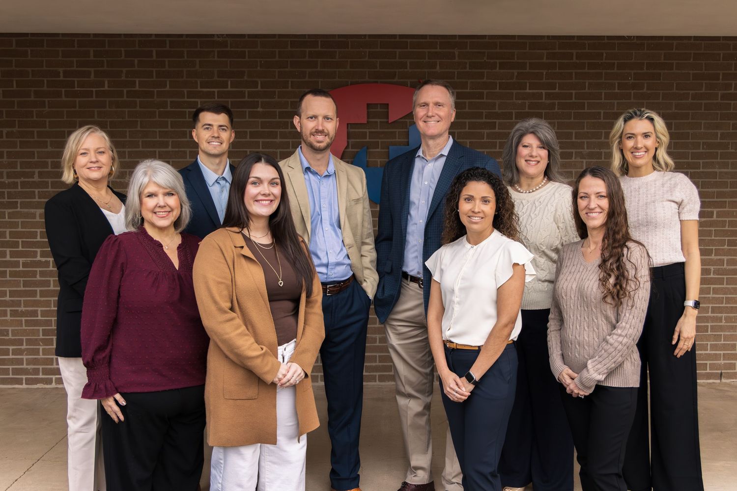 Group of ten smiling professionals, men and women, posing together in front of a brick wall in business casual attire.
