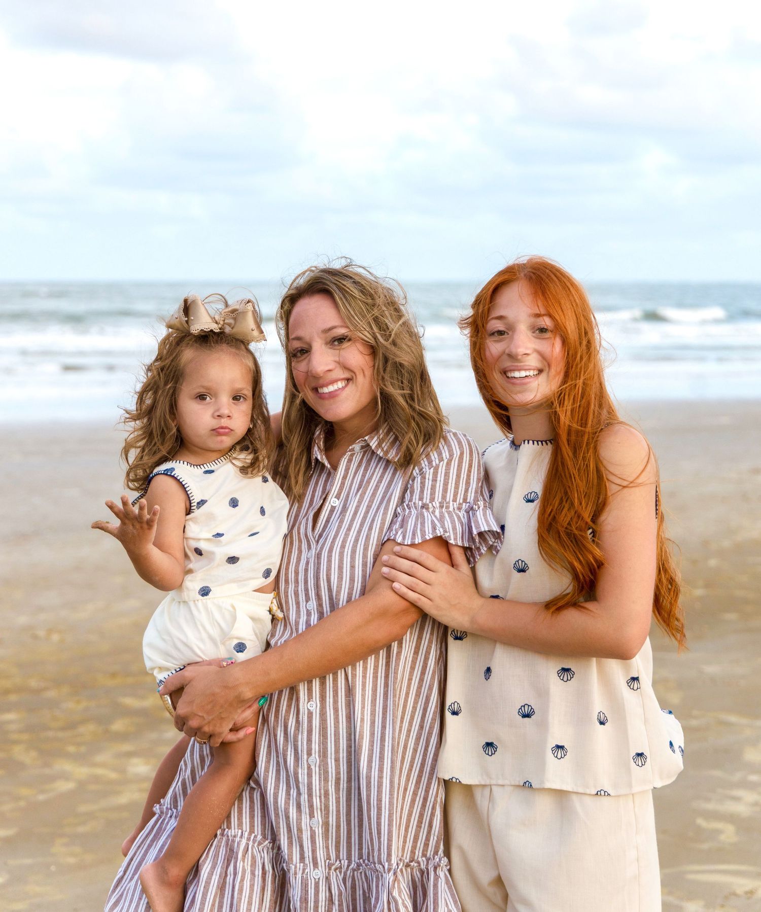 A smiling mother, a red-haired teen girl and a toddler pose together in matching neutral outfits on a sandy beach.