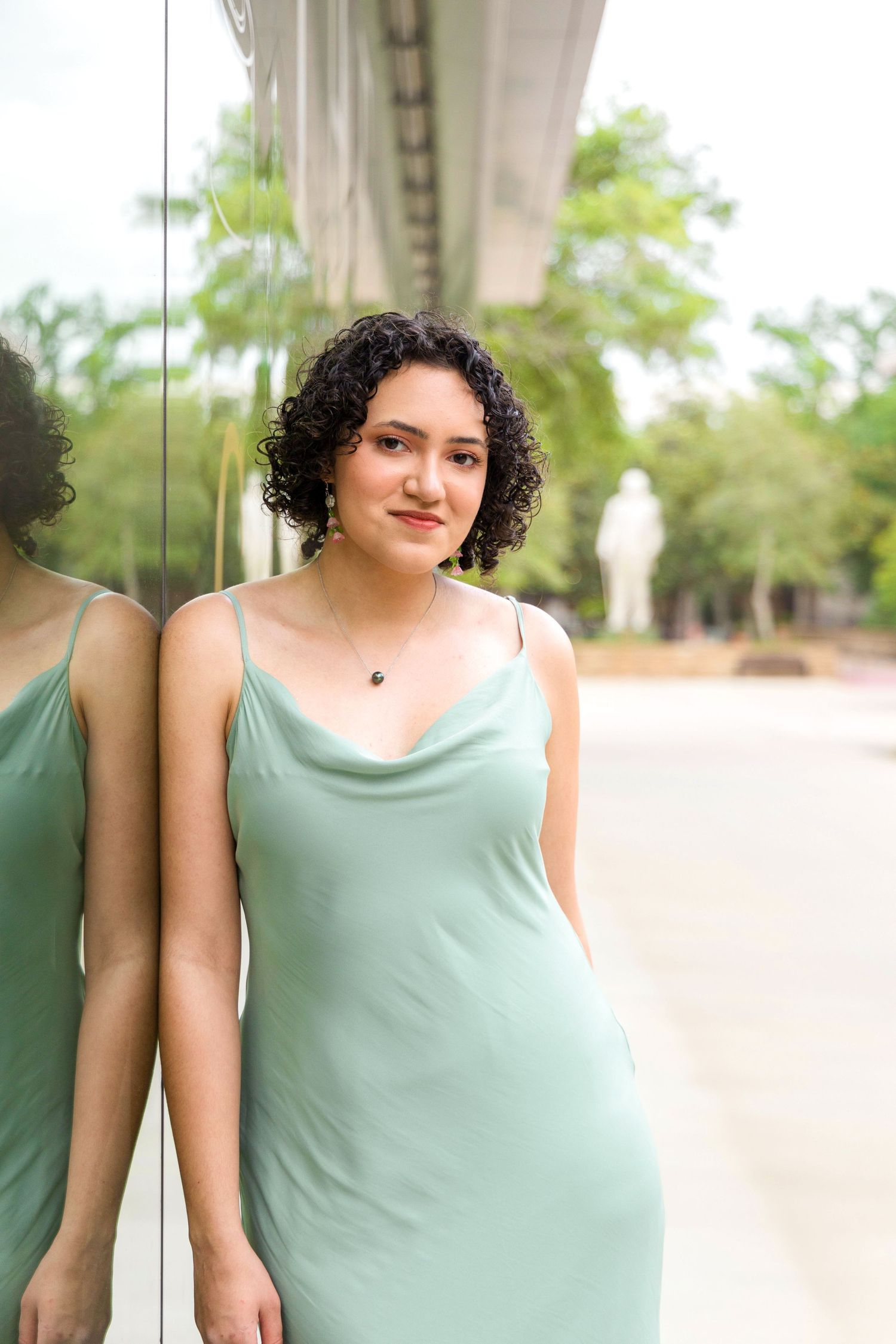 Young woman with dark curly hair in a sage green slip dress leans against a glass wall with her reflection beside her.