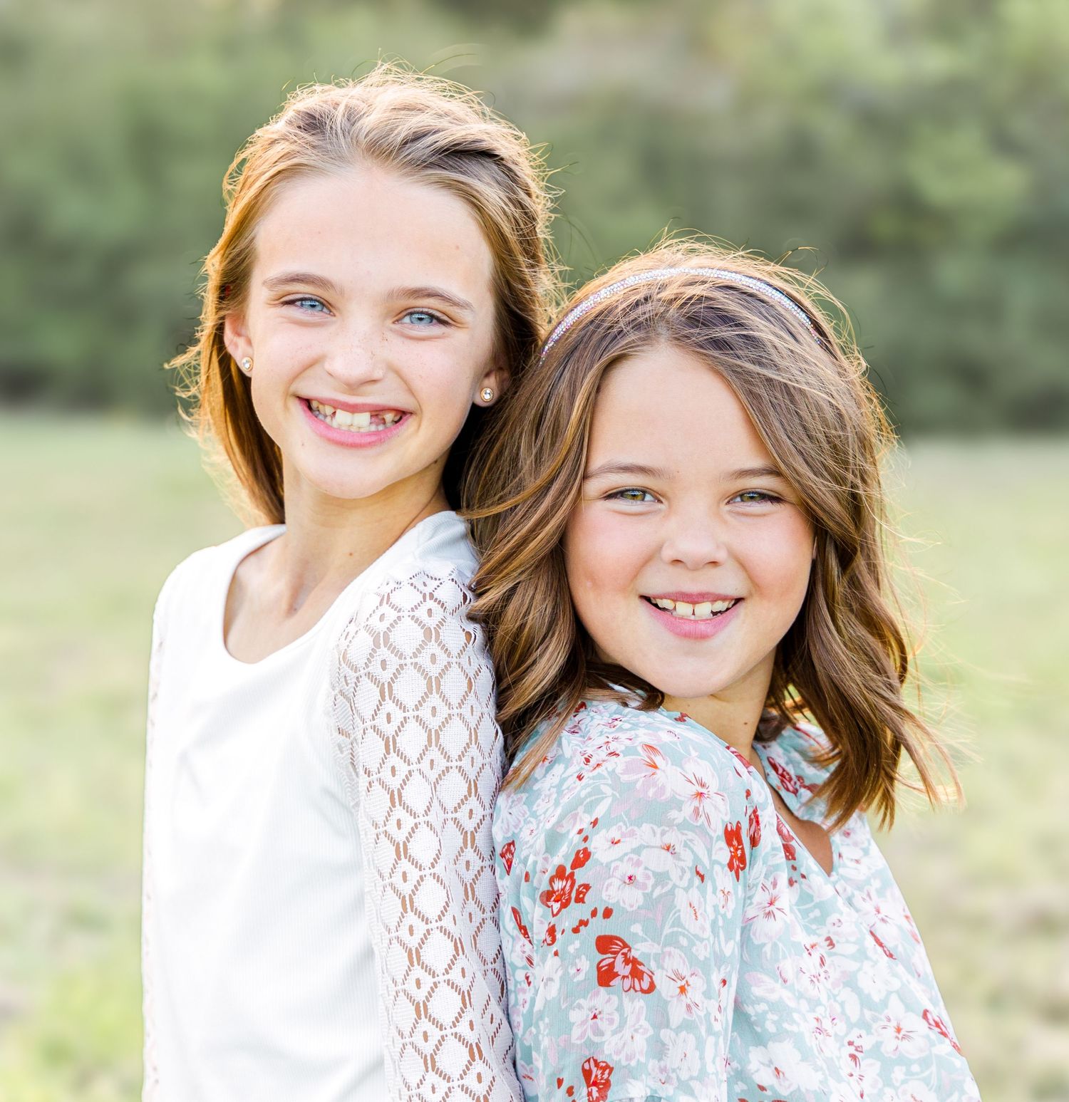 Two smiling young girls posing back to back outdoors in a green field, wearing white lace and floral dresses.