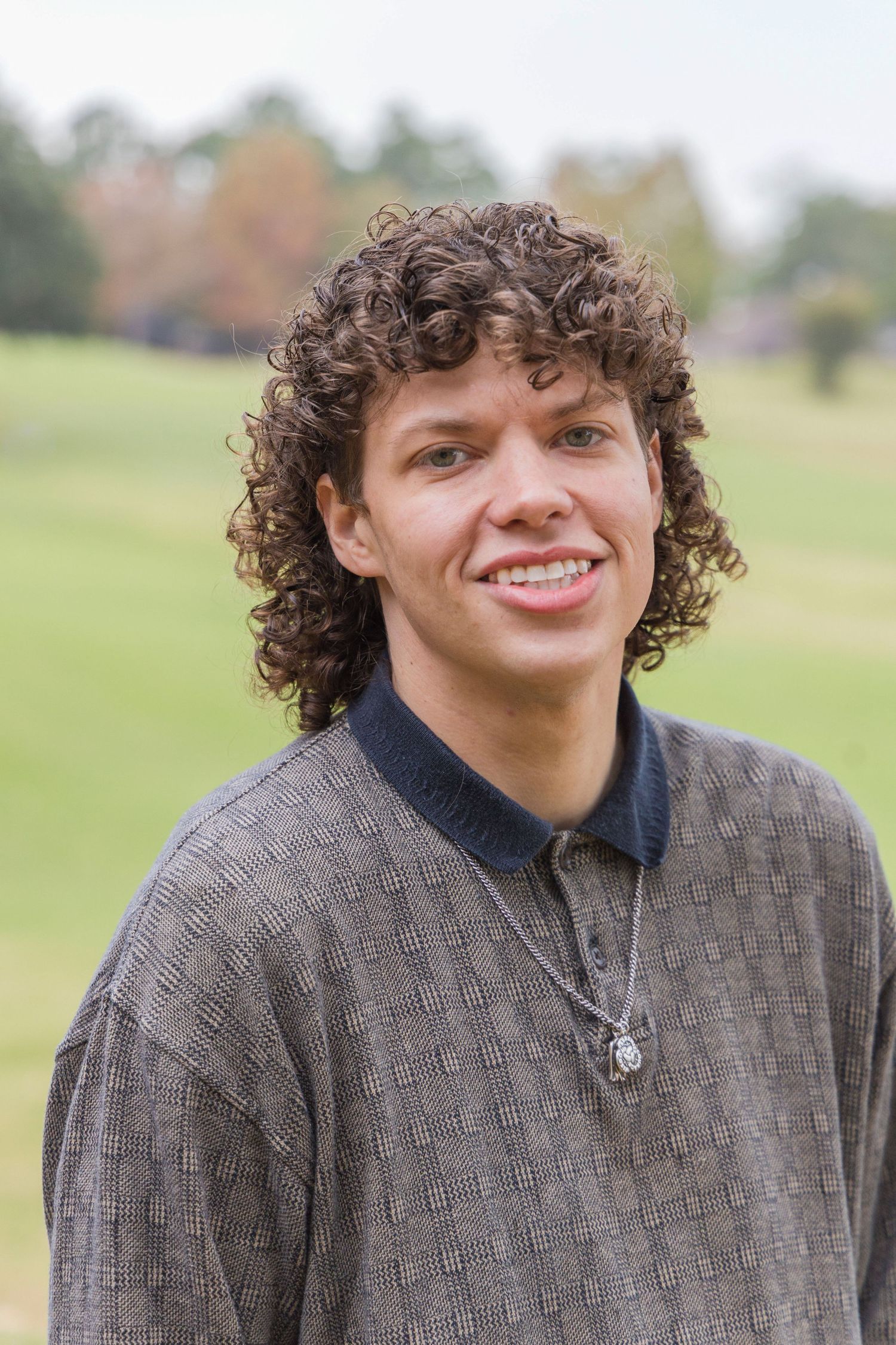 Smiling young man with curly brown hair wearing a gray textured sweater and pendant necklace poses outdoors on a green lawn.