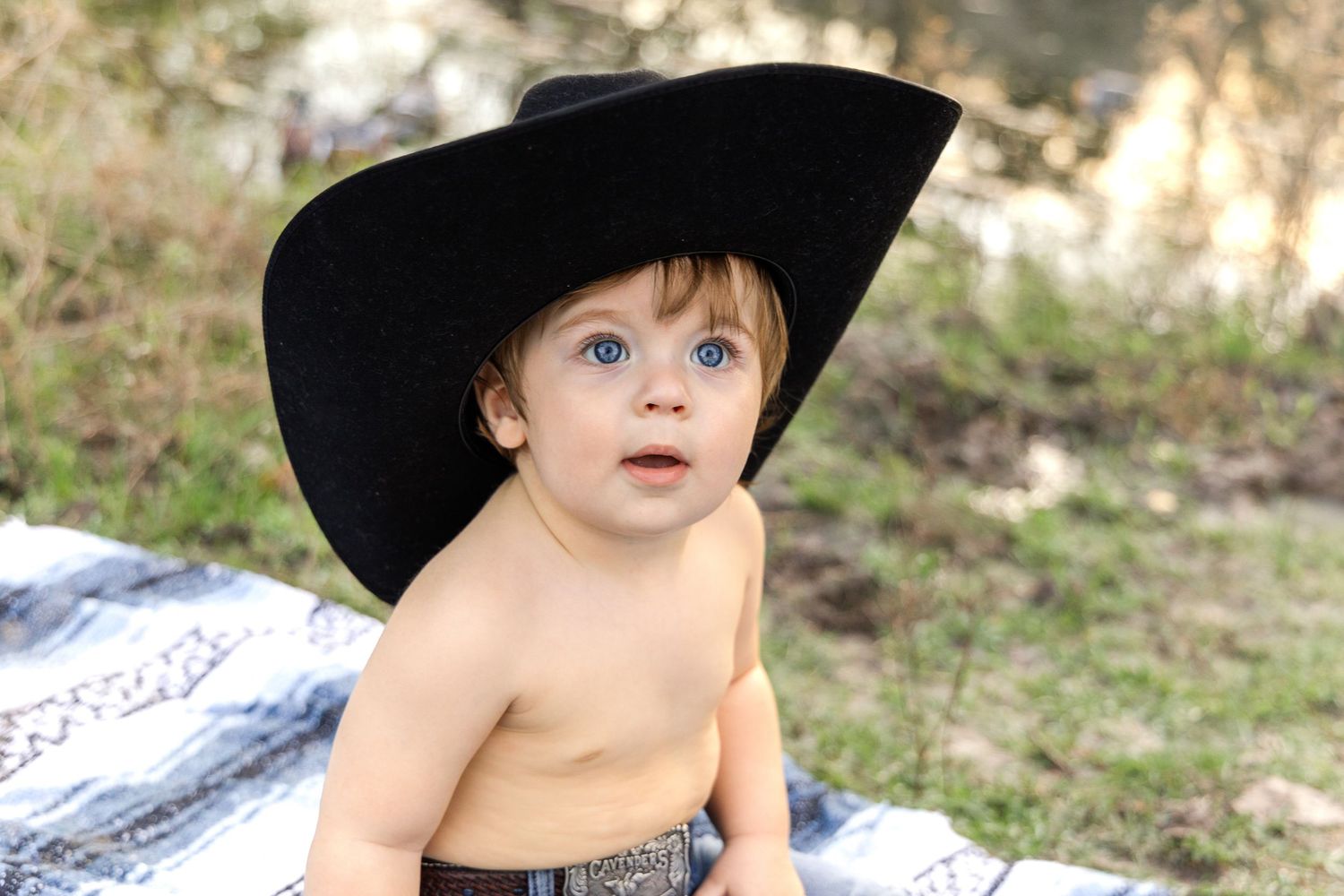 A curious blue-eyed toddler wearing an oversized black cowboy hat sits on a blanket in a grassy outdoor setting.