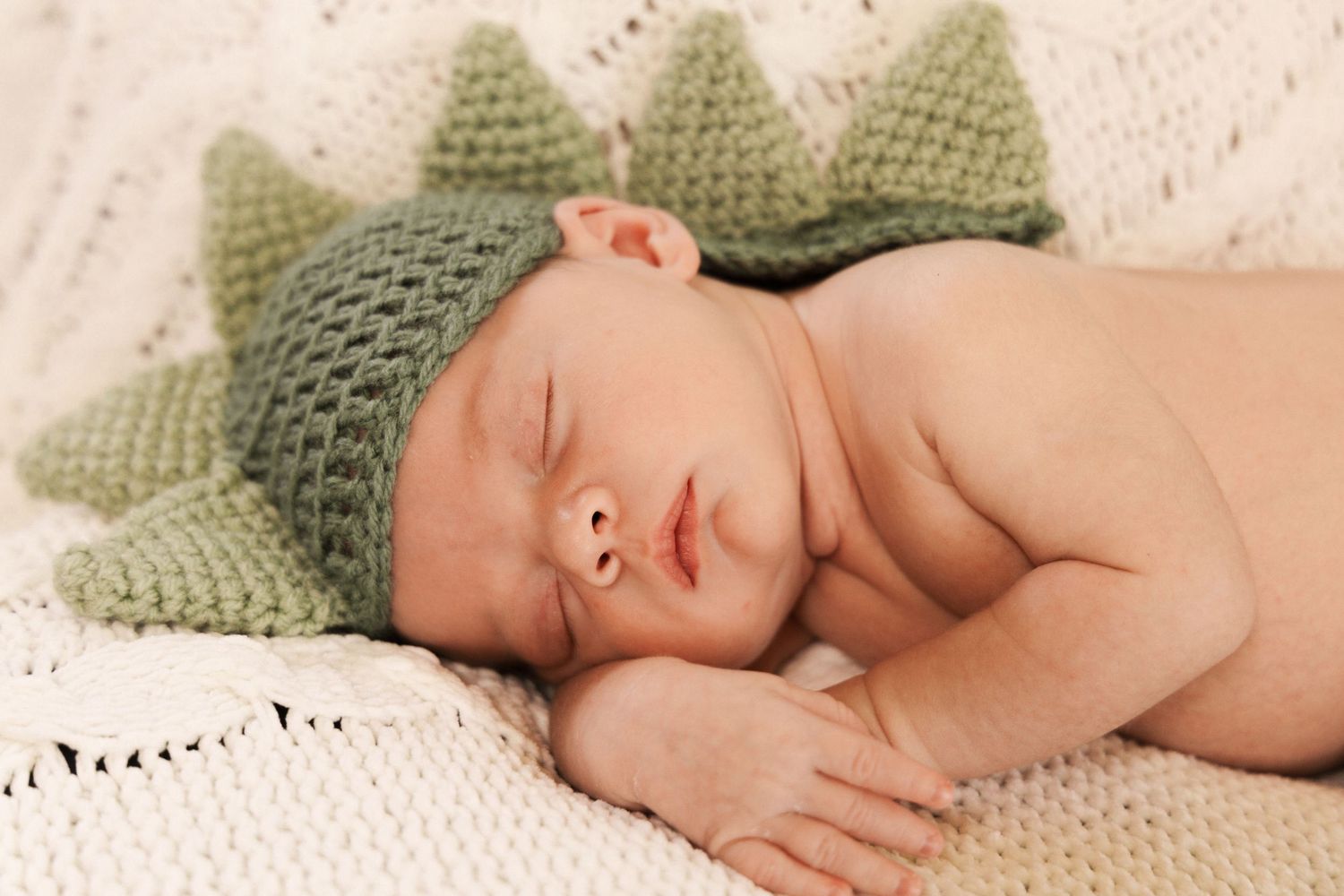Newborn baby sleeping peacefully on a cream knit blanket wearing a green crocheted dinosaur hat with spikes.