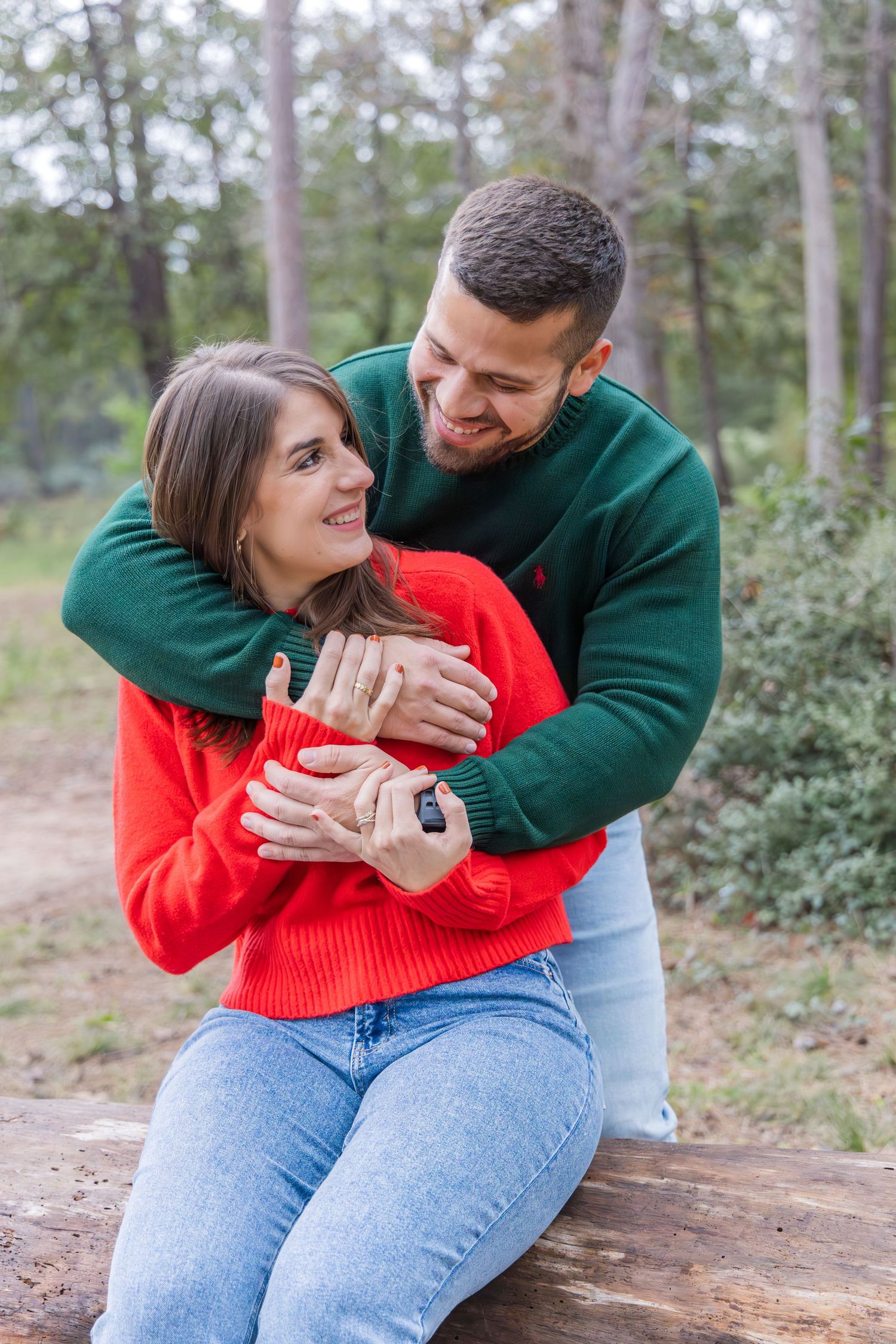 Happy couple in red and green sweaters embrace while sitting on a log in a sunlit forest during an engagement photo session.