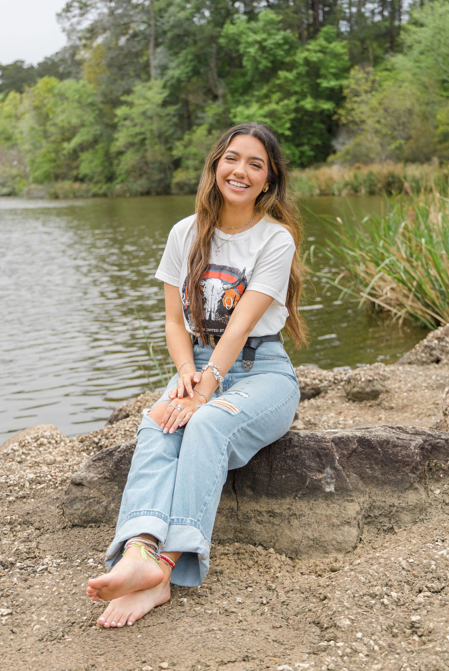 Smiling barefoot young woman in a graphic tee and light-wash jeans sits on a rock beside a calm lake surrounded by trees.