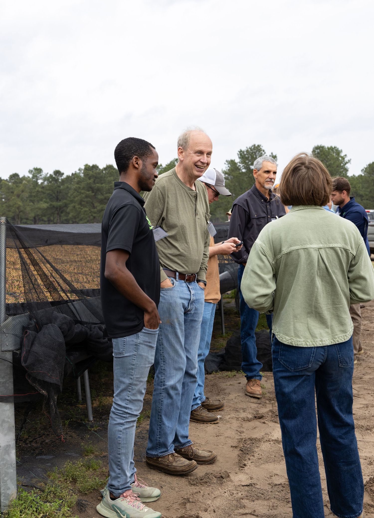 A group of men and women stand talking outdoors on a dirt path near a black shade netting structure, with trees in the background.