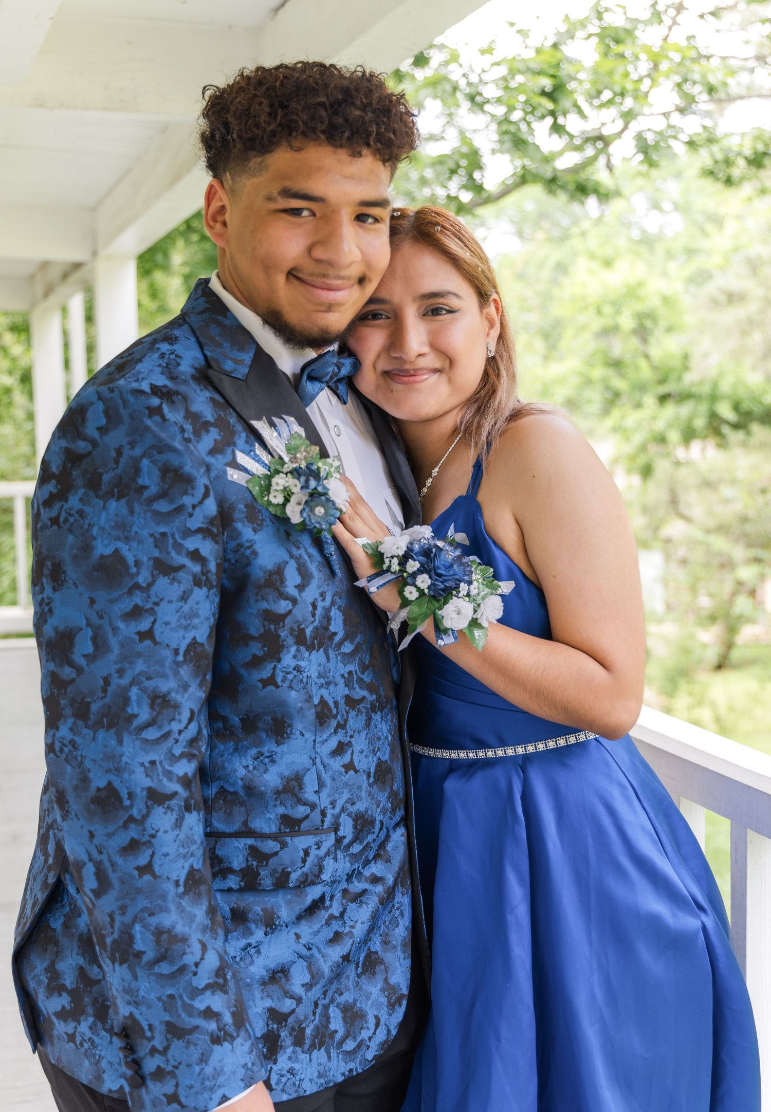 Teen couple in matching blue formal wear with floral corsage and boutonniere smiling together on a porch before prom.