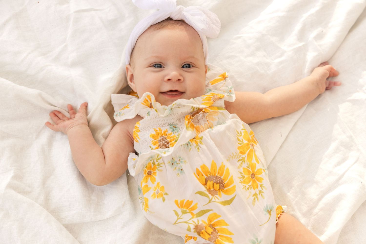 A smiling baby girl in a yellow sunflower romper and white bow headband lies on soft white bedding.