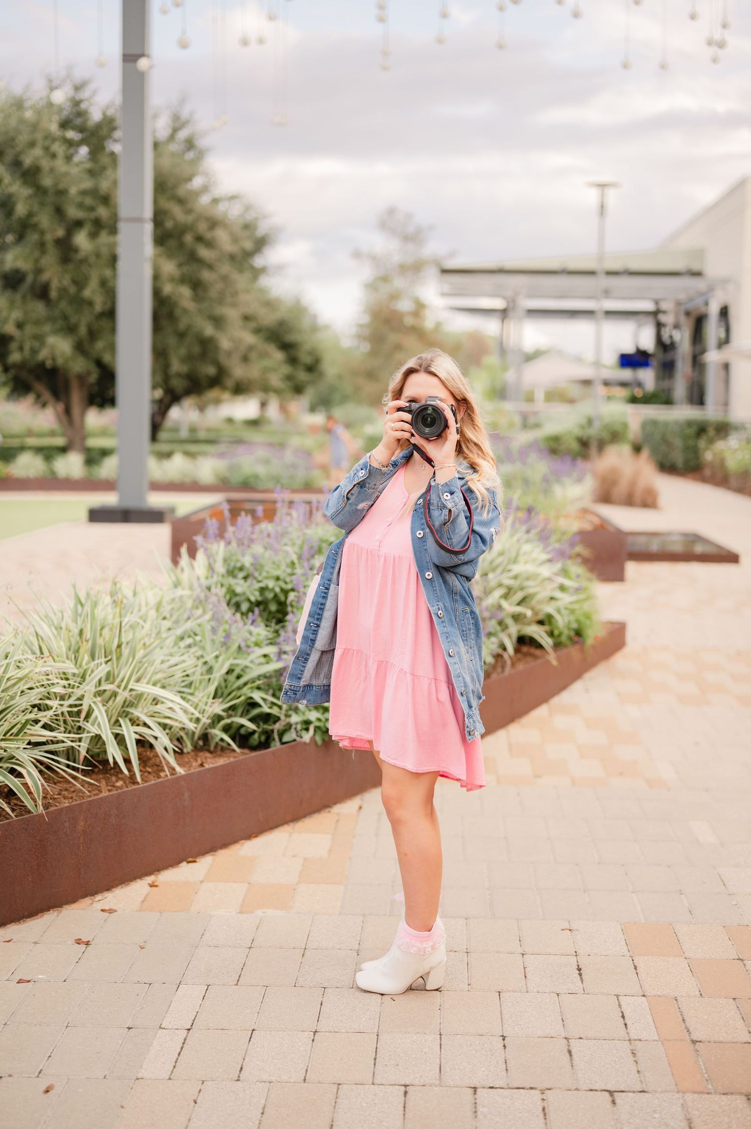 Woman in a pink dress and denim jacket takes a photo with a camera on a brick walkway beside landscaped planters.