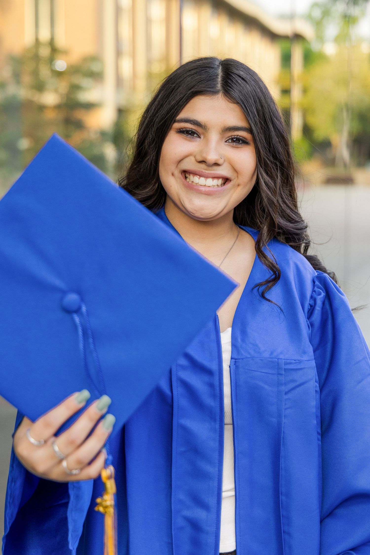 Smiling graduate in a royal blue gown holds a matching mortarboard cap with a gold tassel toward the camera.