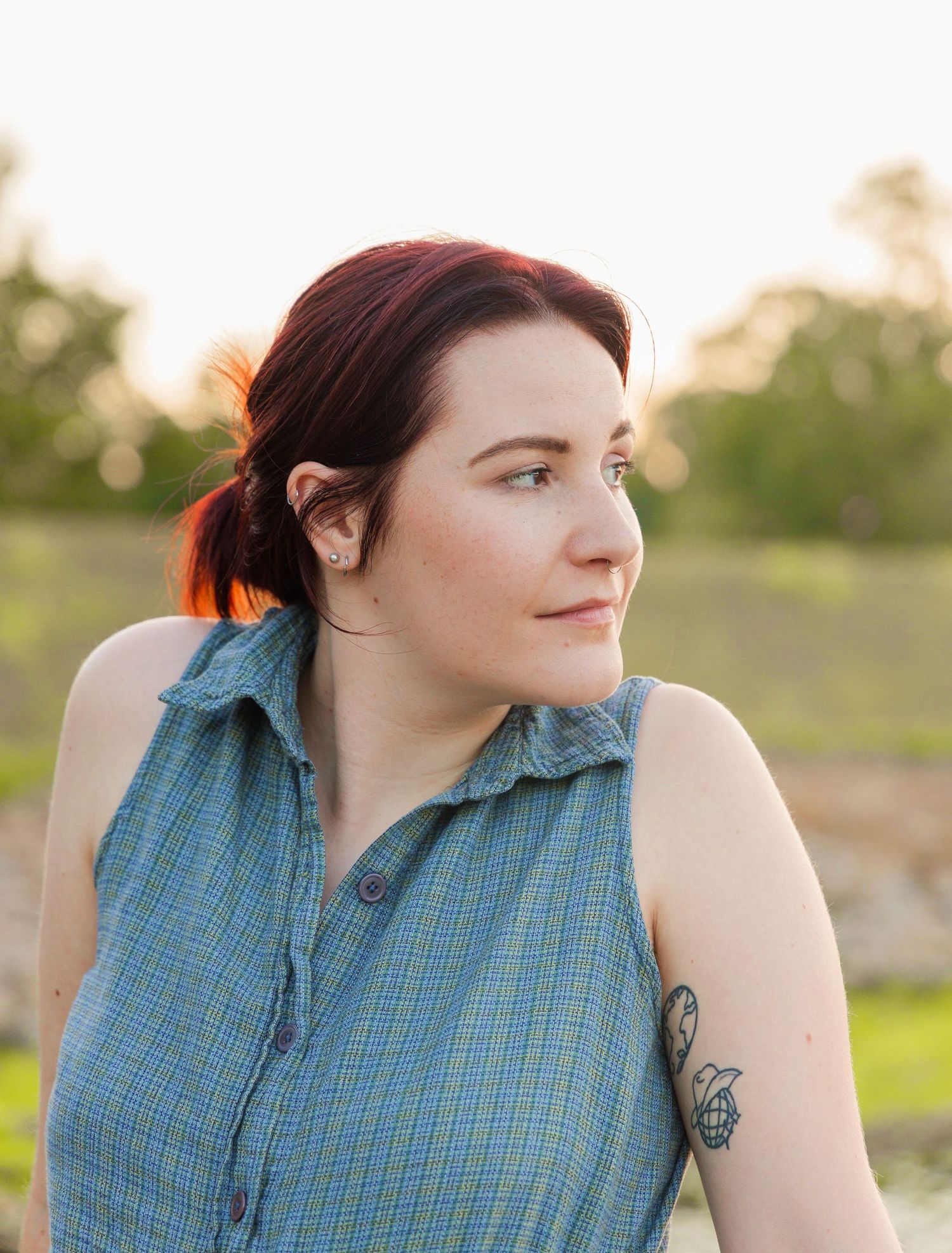 Woman with auburn hair pulled back and an arm tattoo, wearing a sleeveless blue plaid shirt, gazes sideways in warm sunset light.