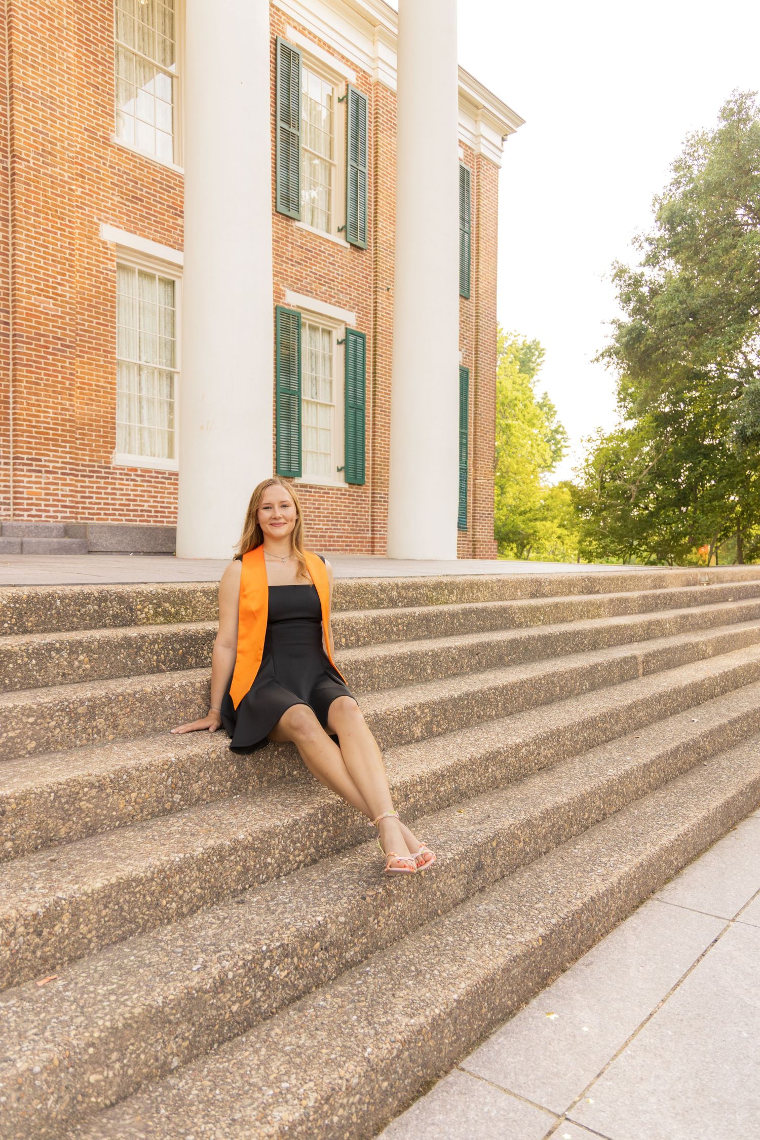 Graduate in a black dress and orange stole sits barefoot on wide stone steps in front of a brick building with white columns.