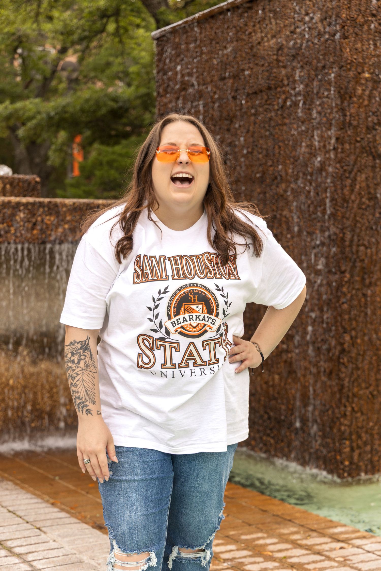 Laughing woman in orange sunglasses, a Sam Houston State University Bearkats shirt and ripped jeans stands in front of a waterfall wall.