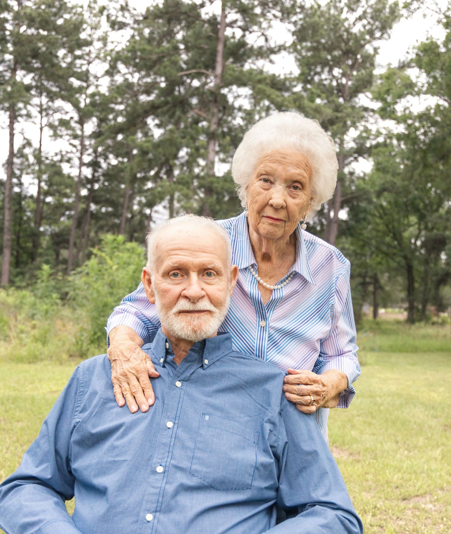 An elderly couple poses lovingly outdoors, the white-haired woman resting her arms on the shoulders of a bearded man in blue.