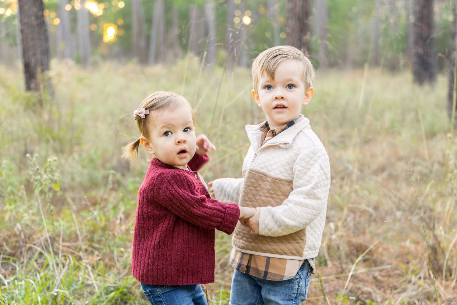 A toddler girl in a maroon sweater holds hands with a young boy in a cream knit sweater in a sunlit autumn forest.