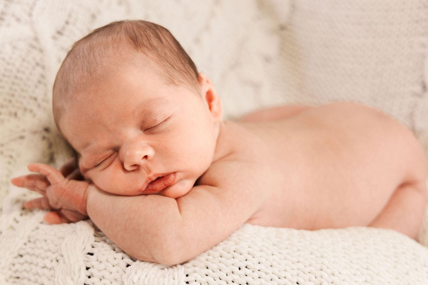 Newborn baby sleeping on its tummy with hands tucked under its chin on a soft cream knit blanket.