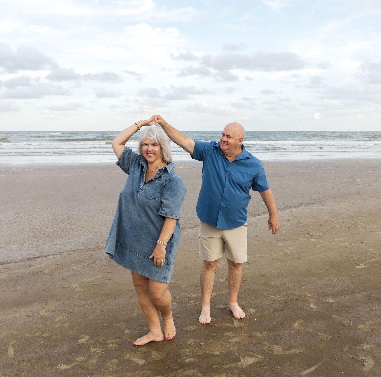 Older couple dancing barefoot on a sandy beach with ocean waves and a cloudy sky in the background.