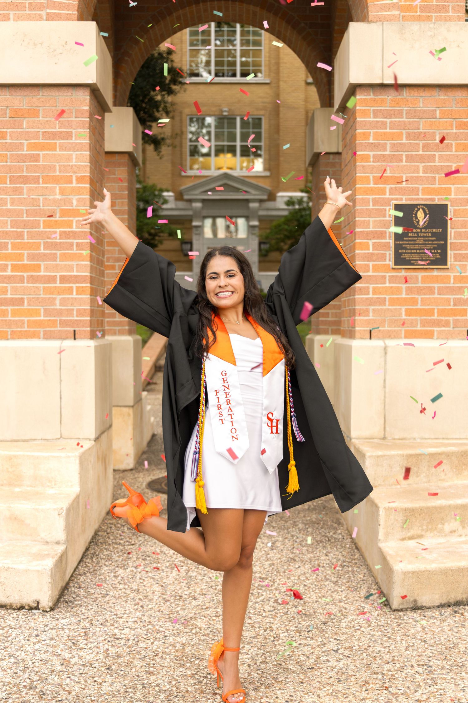 Graduate in a black gown and orange stole leaps joyfully under a brick archway as confetti falls around her.