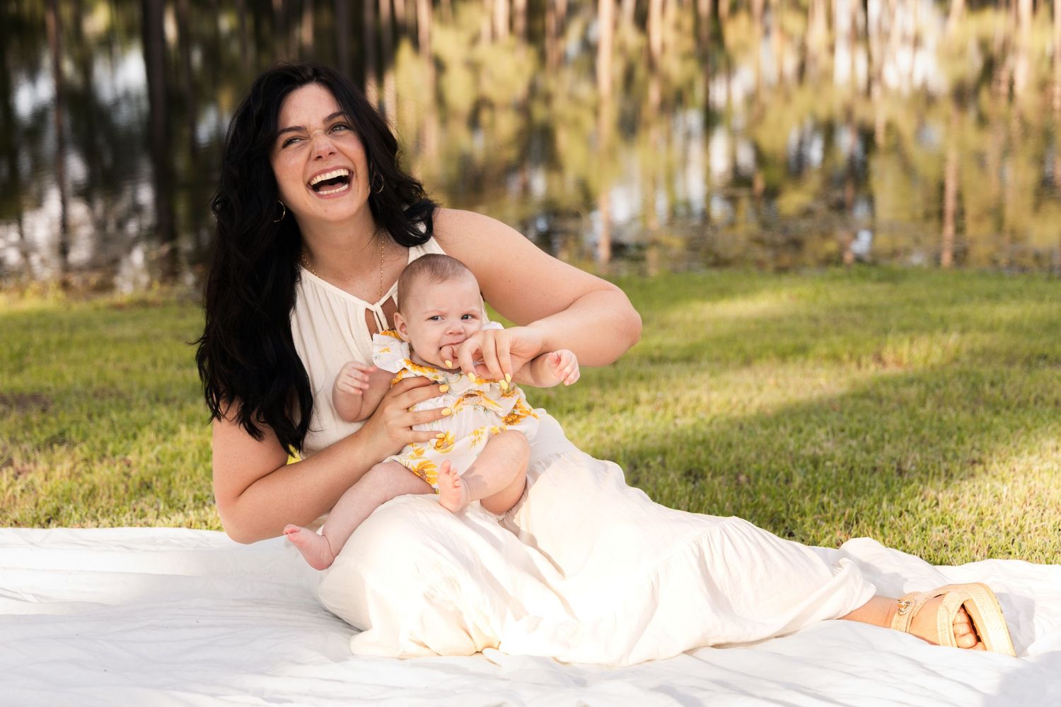 Laughing mother in a white dress holding her baby on a white blanket in the grass beside a reflective lake.