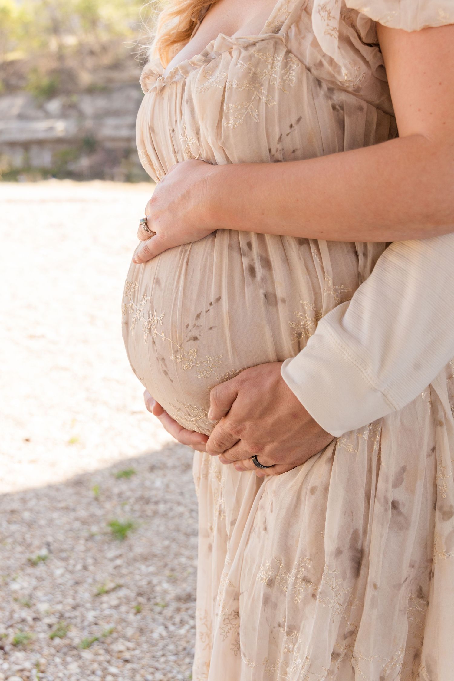 Close-up of an expecting mother in a flowing cream floral dress cradling her baby bump with a partner's hands resting on it.