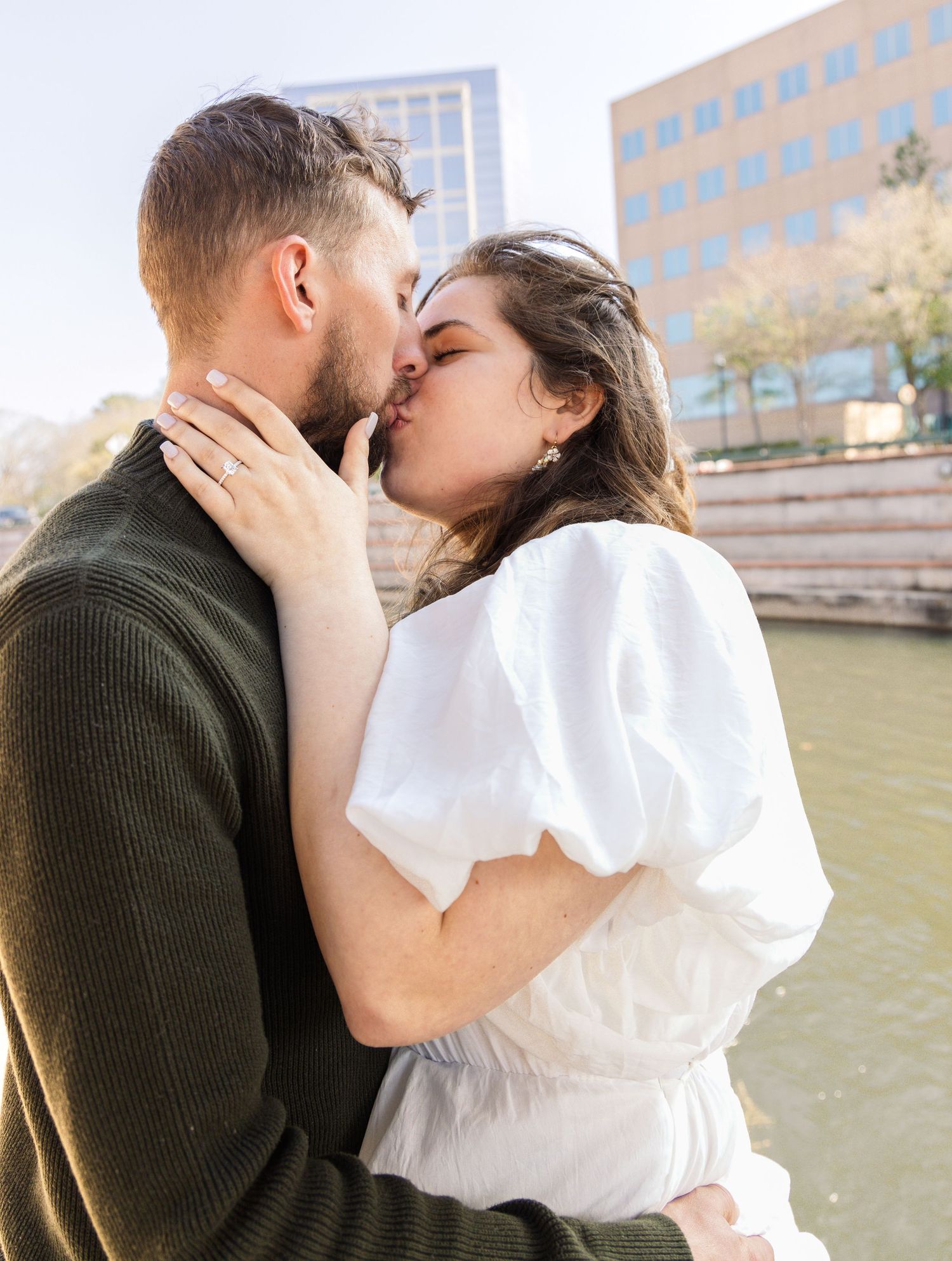 Couple sharing a kiss beside a riverside while the woman's hand displays her engagement ring in a city setting.