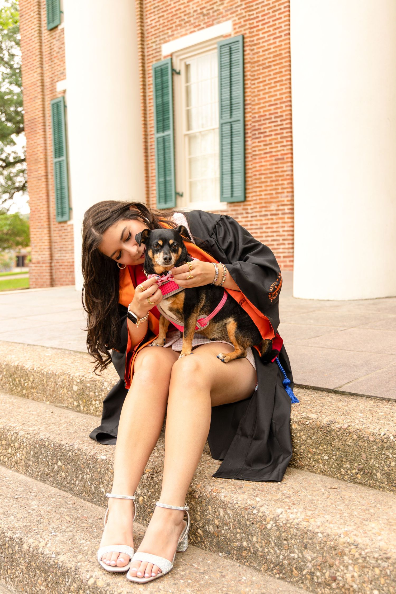 Graduate in a black gown and orange stole sits on brick steps outside a columned building, hugging a small dog.
